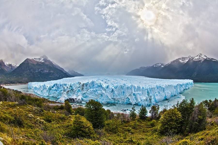 Panoramic view of the Glaciar Perito Moreno, white blue ice, turquoise colored lake, dark gray stormy sky, forest and mountains